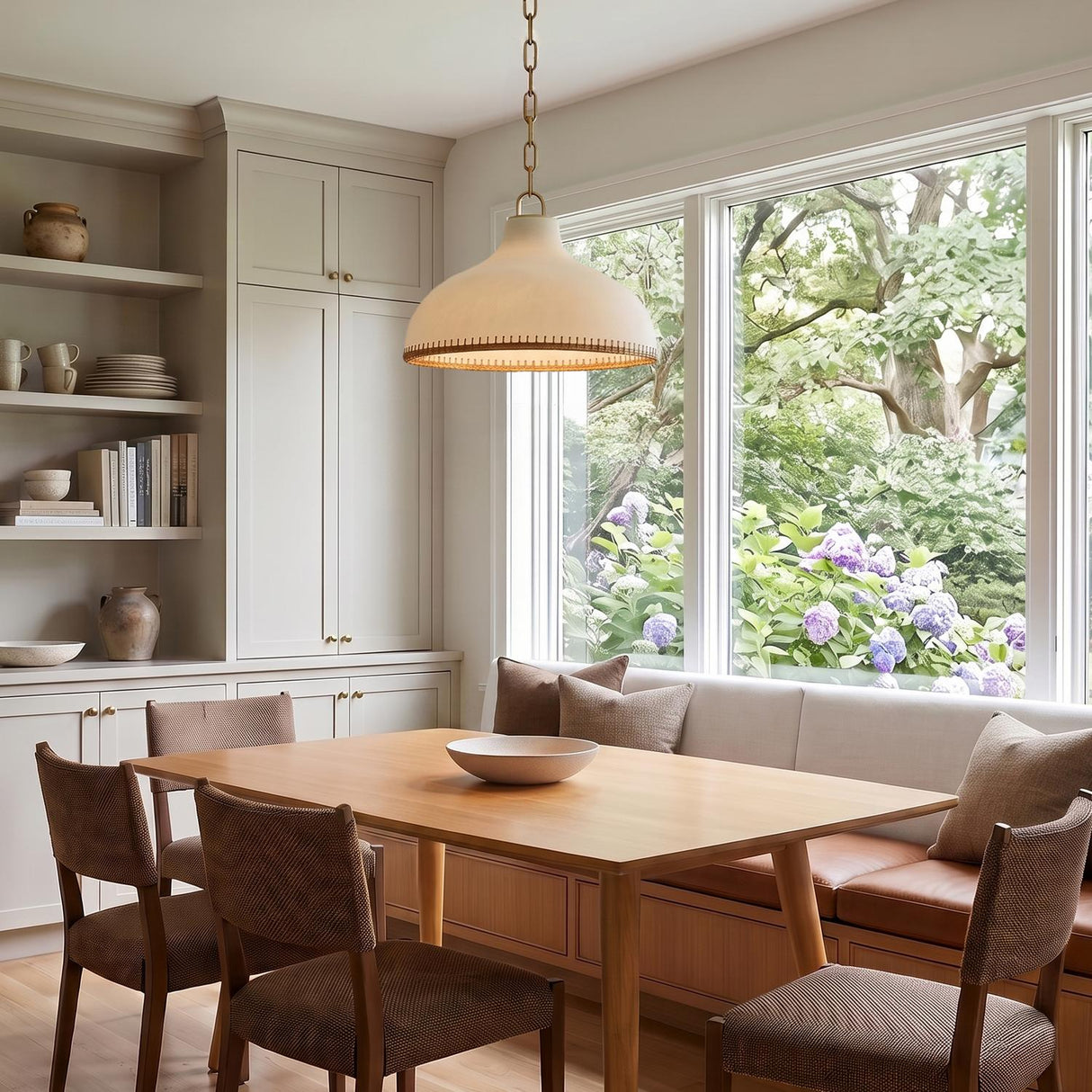 A transitional dining room featuring a cream and rattan pendant with aged brass details over a wood table beside built-in cabinets and large windows.