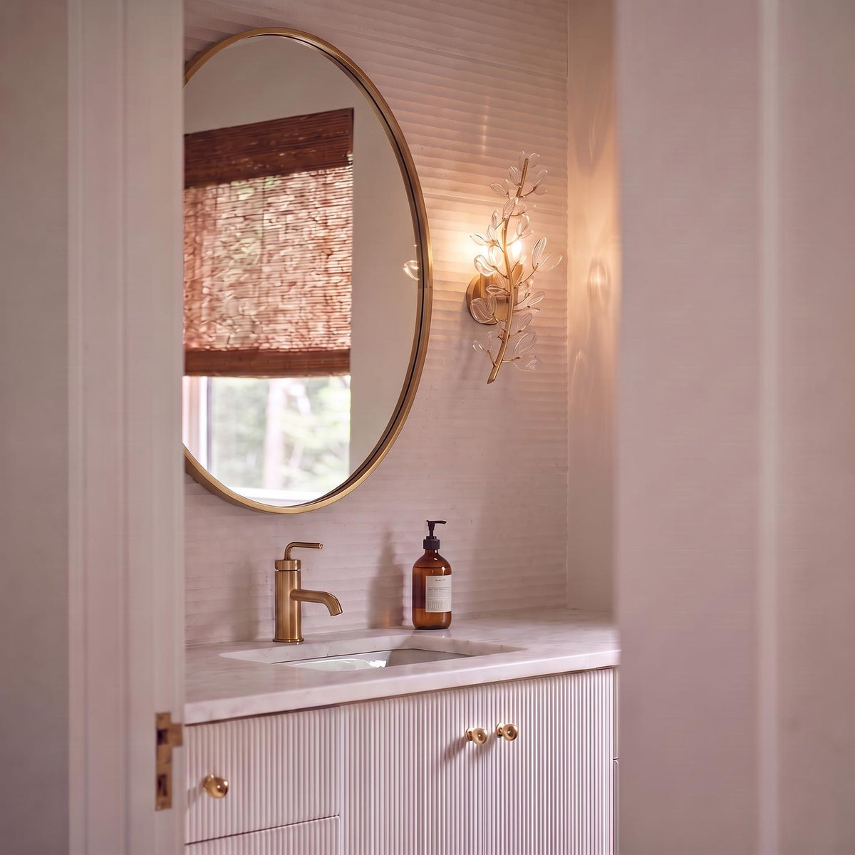 A contemporary bathroom featuring the Esme Wall Sconce 19" in soft brass and clear glass mounted beside a circular gold mirror above a white marble vanity.