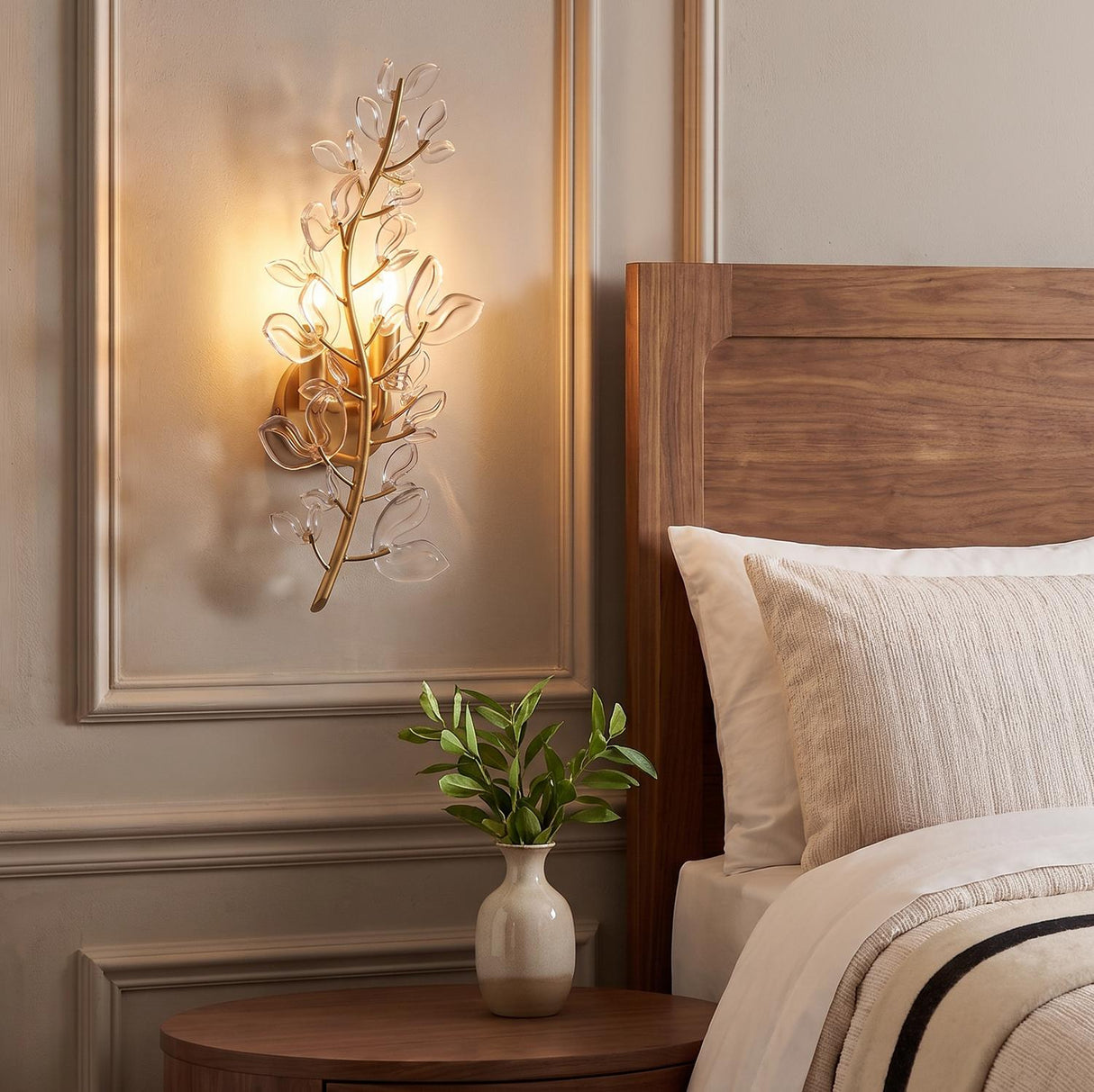 A contemporary bedroom with a soft brass and clear glass wall sconce mounted on beige paneled walls beside a warm walnut headboard and nightstand.