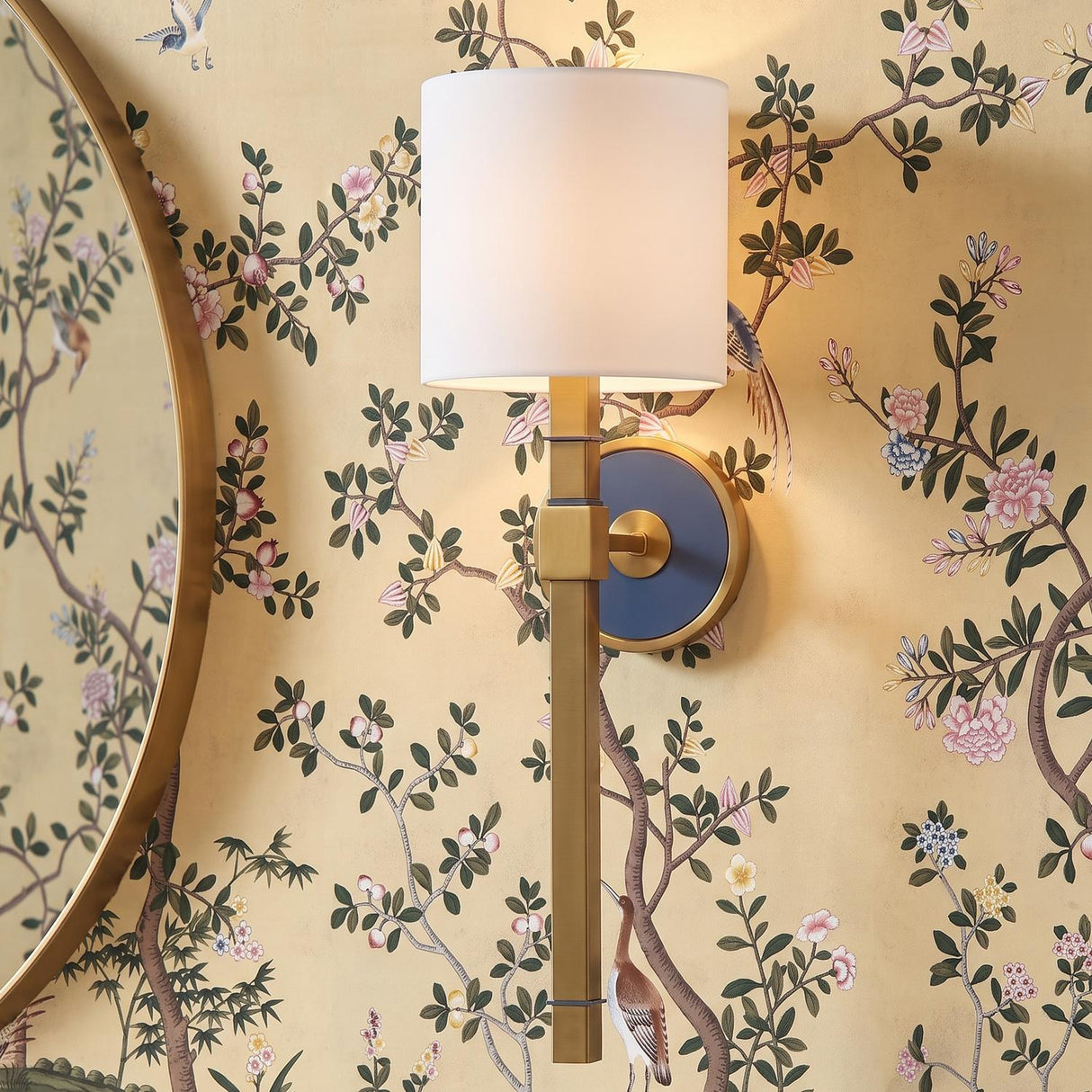 A traditional foyer with a Ramona Wall Sconce 21", Brass and Vintage Blue with White Shade mounted on floral chinoiserie wallpaper beside a circular gold mirror.
