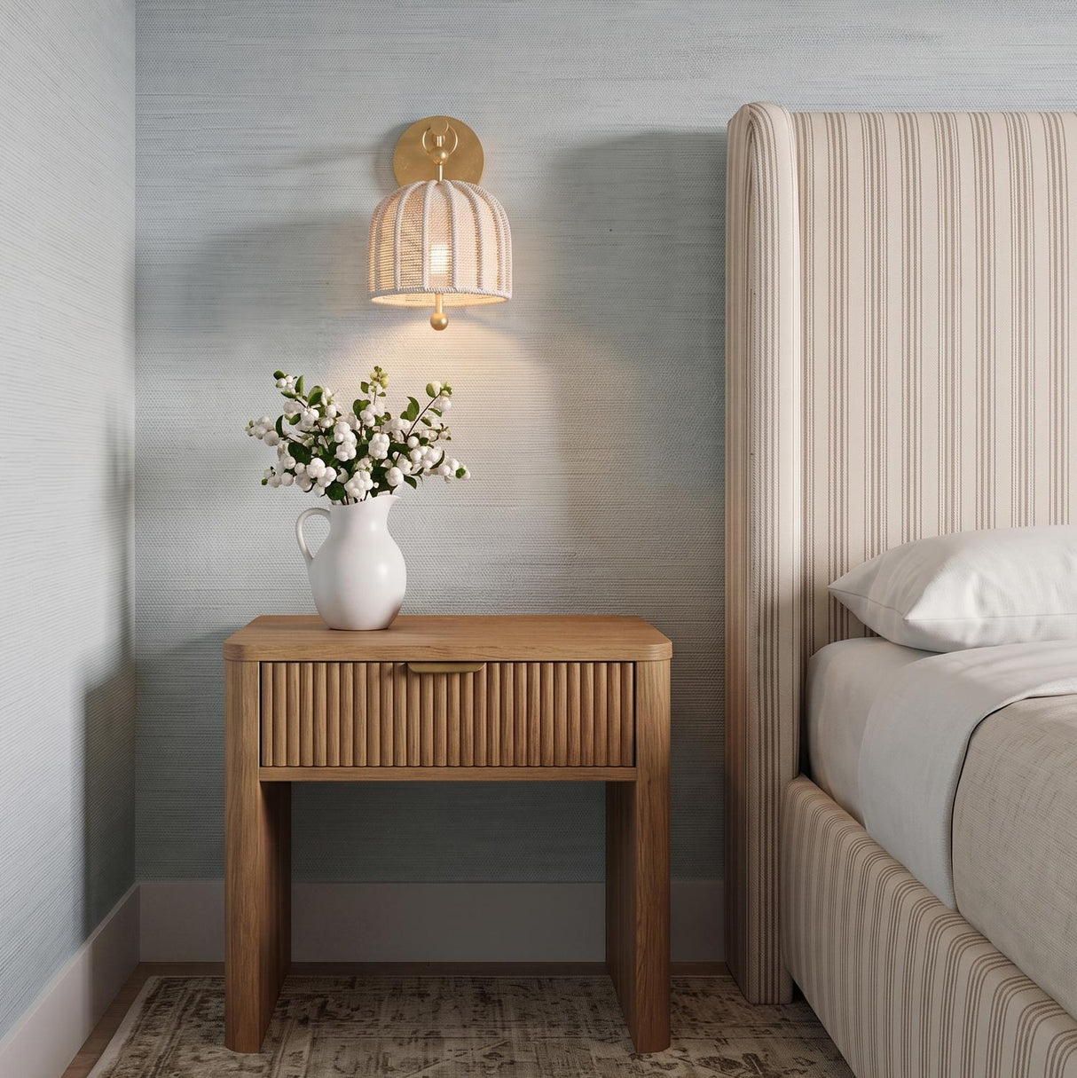 A coastal bedroom featuring the Benito Wall Sconce 12 inch in gold leaf and natural rope mounted above a fluted wood nightstand and striped headboard.