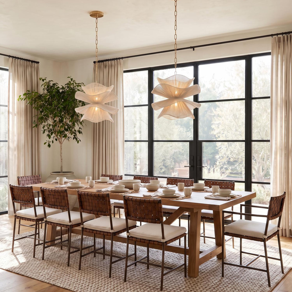 A transitional dining room with two Lotus Pendant 20-inch Aged Brass and Natural Cotton fixtures hanging over a wood table with woven leather chairs and large windows.