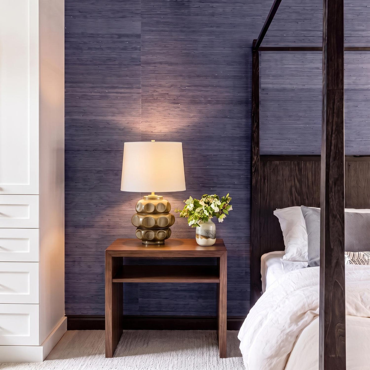 A contemporary bedroom with the Portman Table Lamp 21", Mocha Glaze and Aged Brass on a walnut nightstand against textured purple wallpaper beside a dark wood four-poster bed.