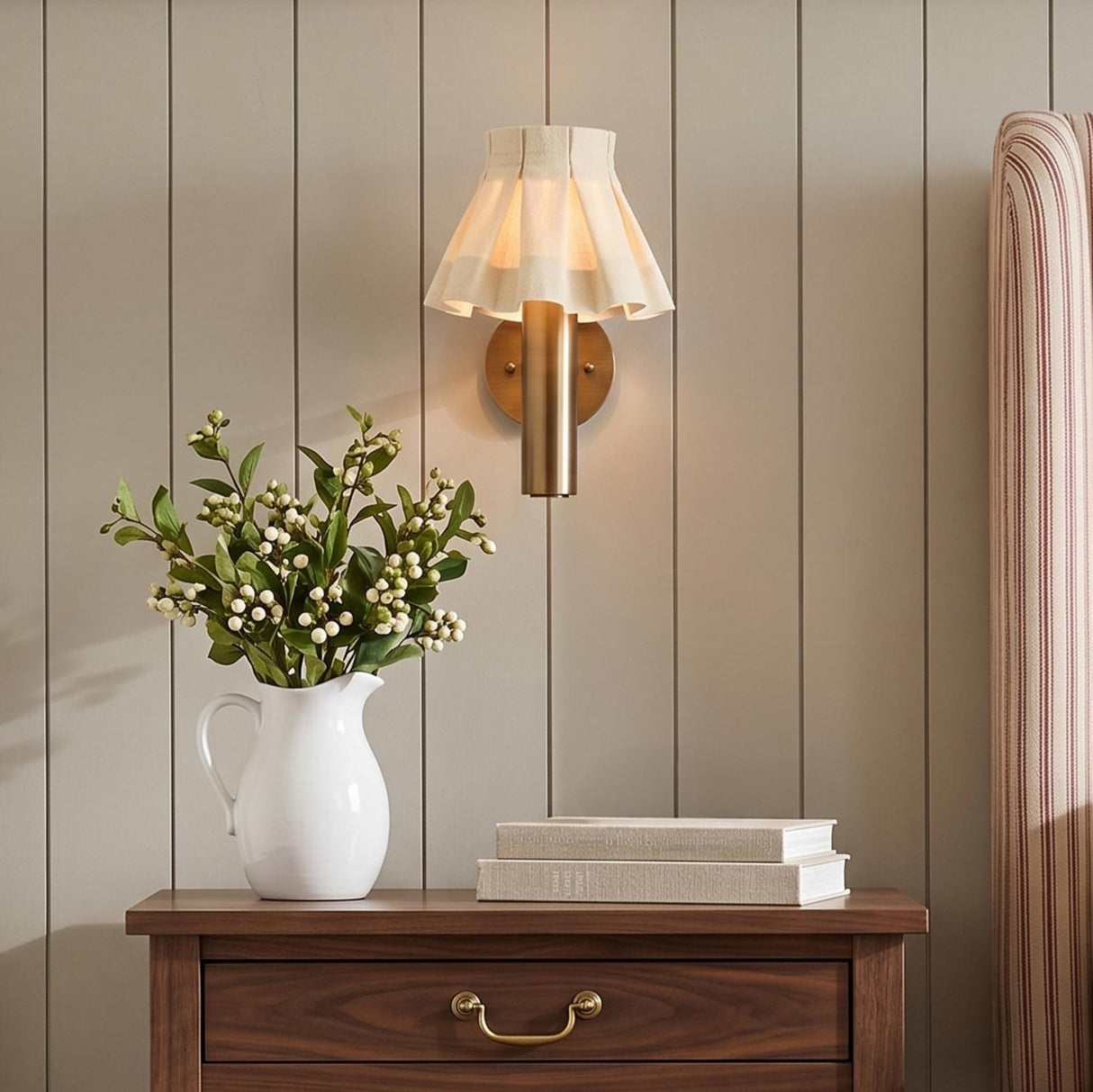A traditional bedroom featuring the Tabitha Wall Sconce in patina brass and wheat pleated linen mounted on a paneled wall above a walnut nightstand.