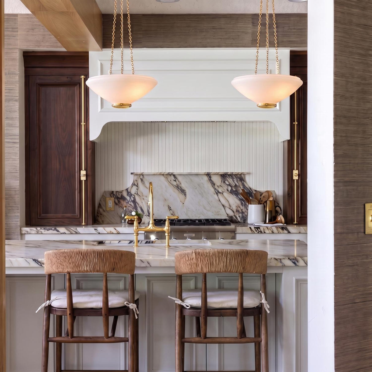 A transitional kitchen featuring two Lindor Medium Pendants in aged brass and opal glass over a marble island with walnut cabinets and woven bar stools.