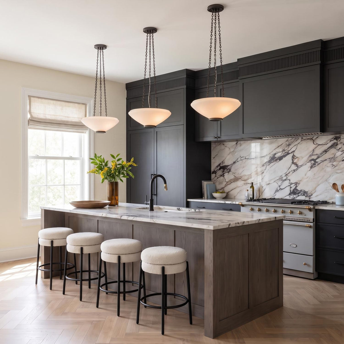 A transitional kitchen featuring three Lindor Medium Pendants in distressed bronze and opal glass suspended over a wood island with marble countertops and black cabinetry.