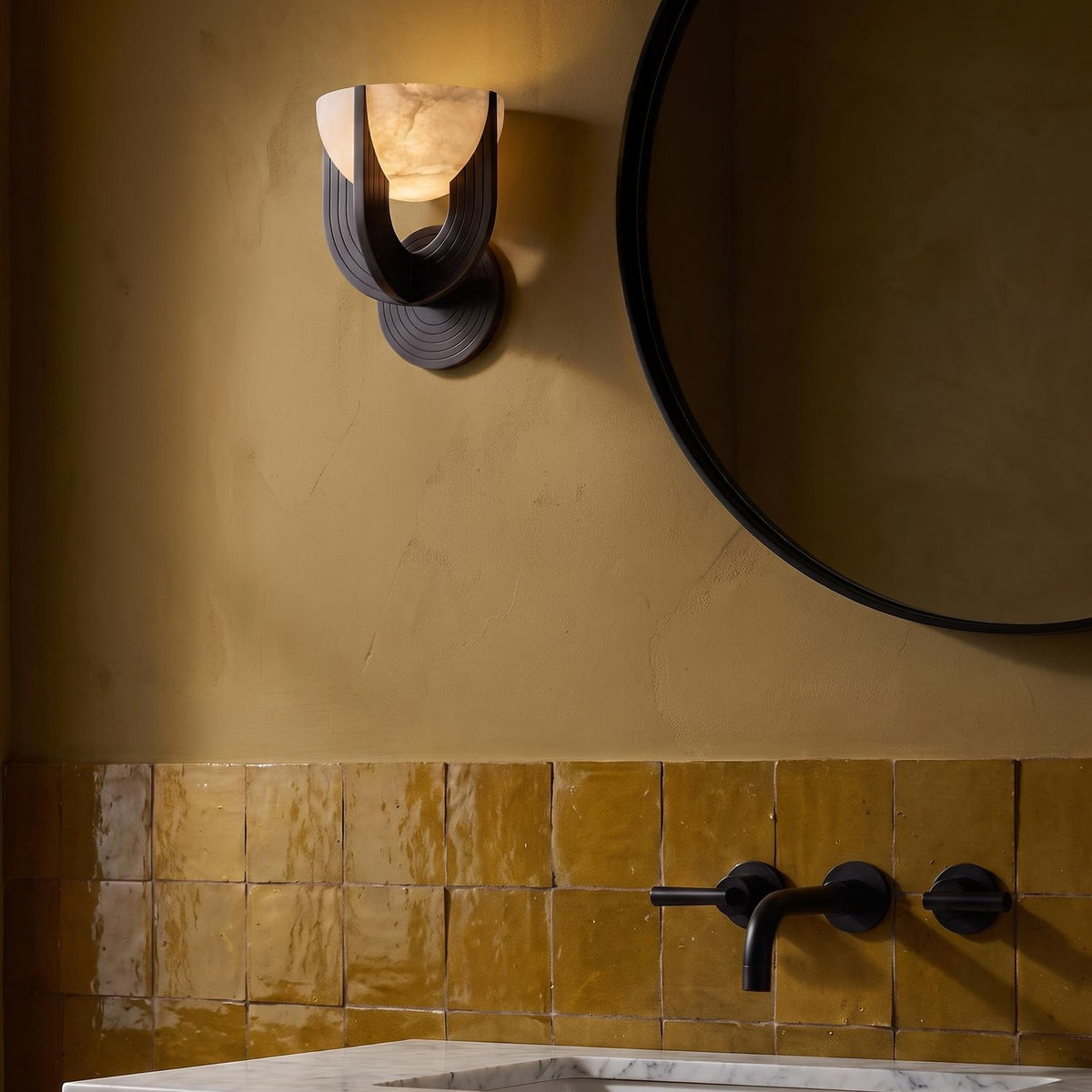 A modern bathroom with an Armand LED Wall Sconce 8", Urban Bronze and Alabaster mounted beside a round black mirror over ochre zellige tile and marble vanity.