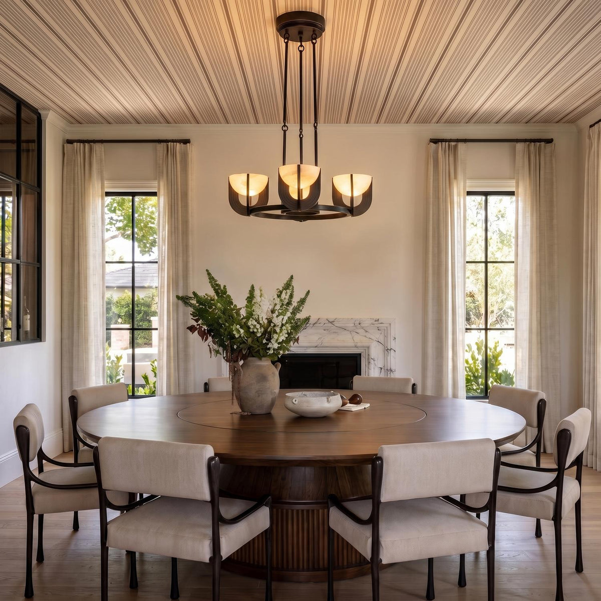 A transitional dining room featuring the Armand LED Chandelier with an urban bronze finish and alabaster shades over a round walnut table and marble fireplace.