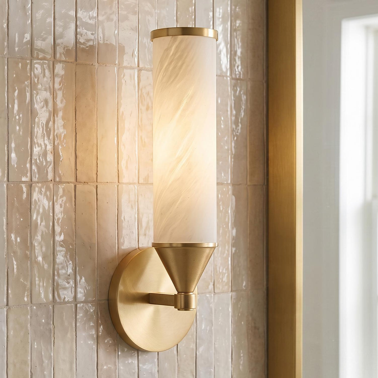 A contemporary bathroom featuring a Sayer Wall Sconce in aged brass and cloud glass mounted on a cream ceramic tile wall beside a gold framed mirror.