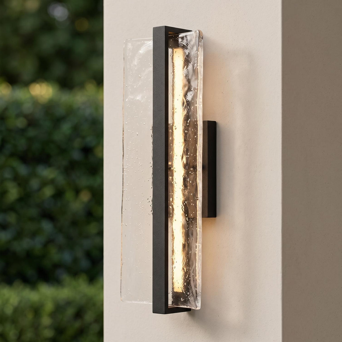A modern patio setting with a Kendrick LED Outdoor Wall Light in textured black and seeded cast glass glowing on a beige stucco pillar beside green garden hedges.