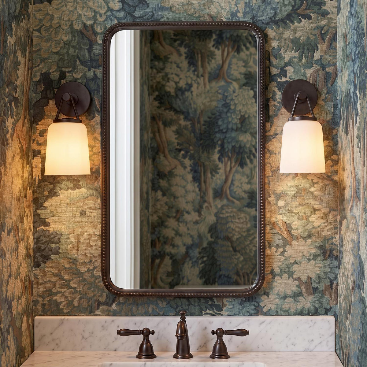 A traditional bathroom with two aged iron and opal glass wall sconces flanking a beaded mirror above a white marble vanity and forest landscape wallpaper.