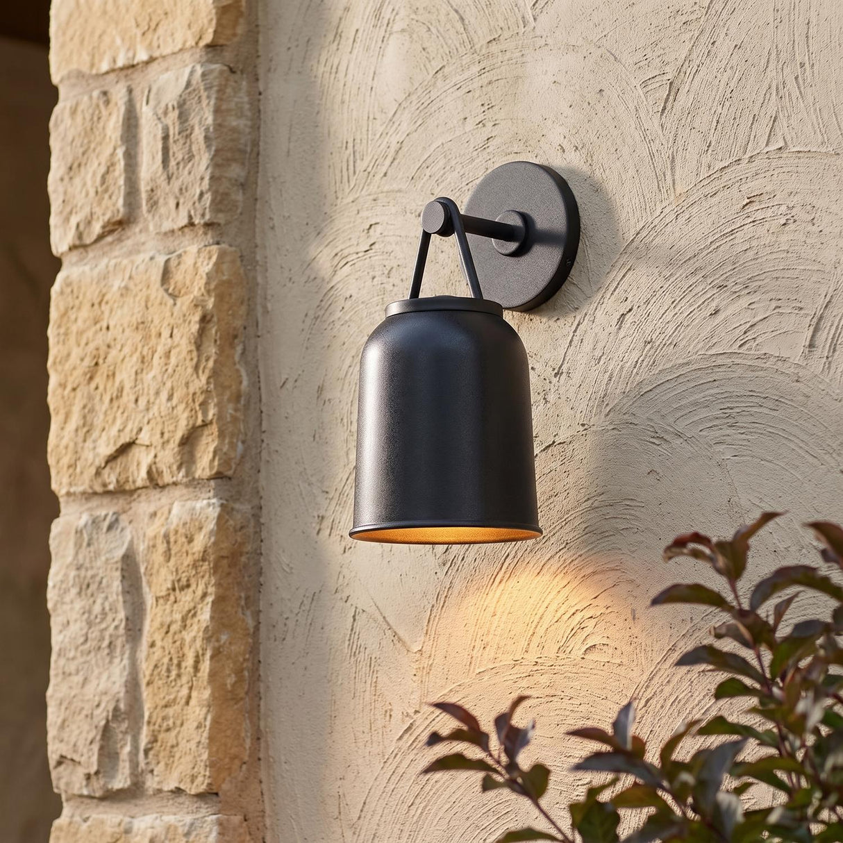 A Mediterranean home exterior with a Stratford Outdoor Wall Sconce, Small, Textured Black glowing on a beige stucco wall beside a stone pillar with dark green foliage.