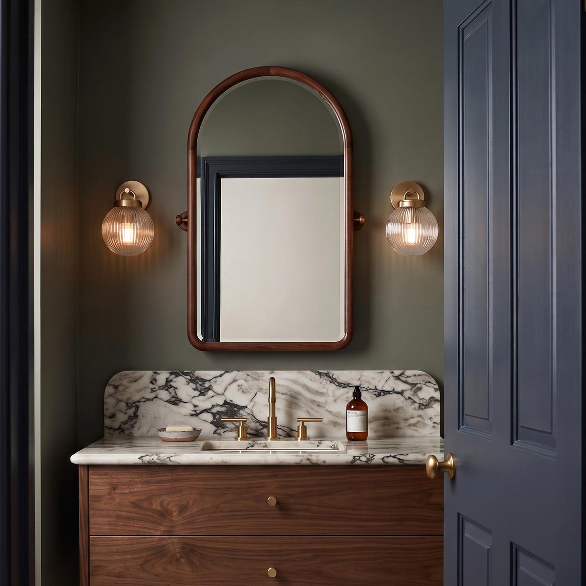 A mid-century bathroom with two Whitaker Wall Sconce 9-inch fixtures in satin brass and clear ribbed glass flanking an arched walnut mirror over a marble backsplash.