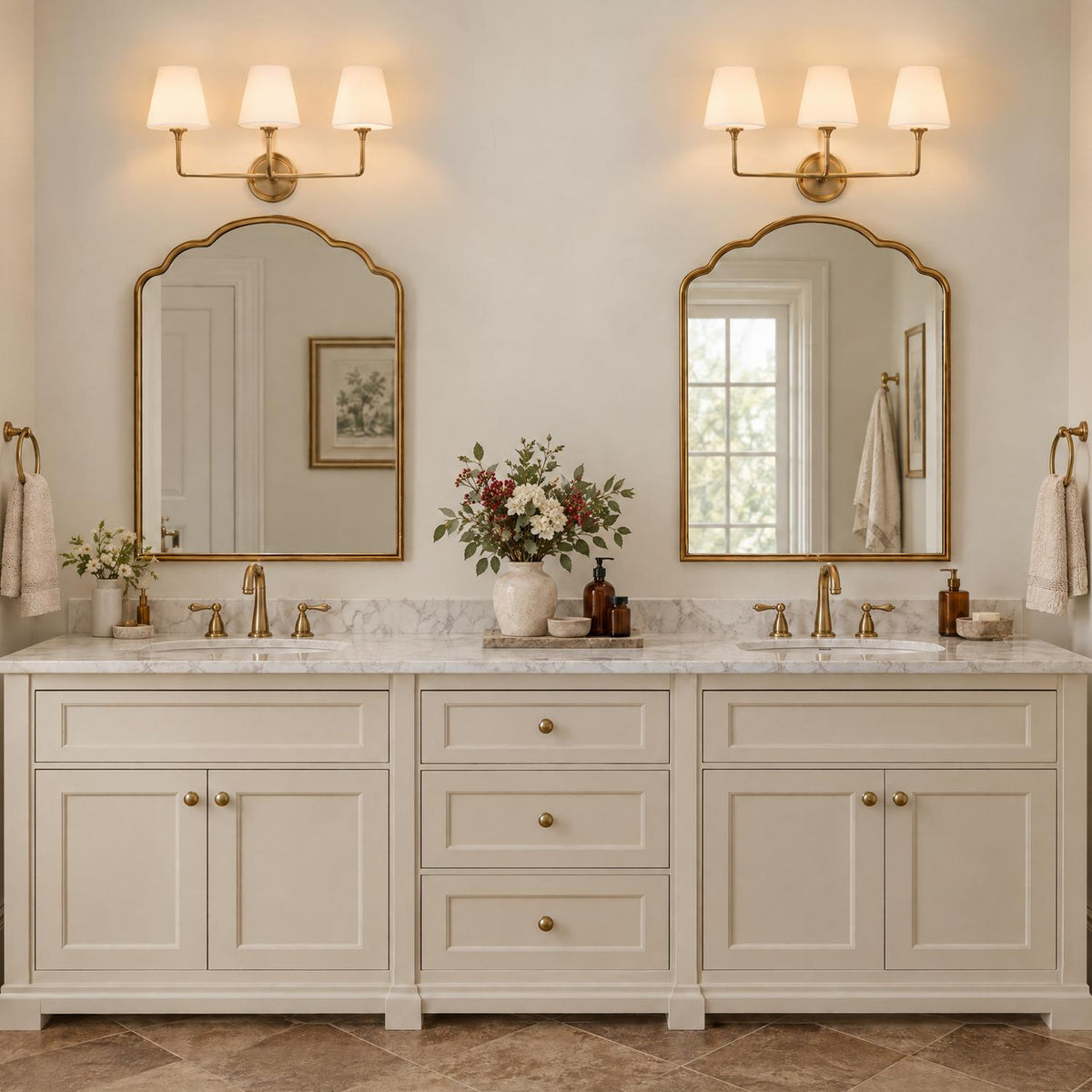 A transitional bathroom features two brushed brass 3-light vanity lights with white fabric shades mounted above arched mirrors over a marble double vanity.