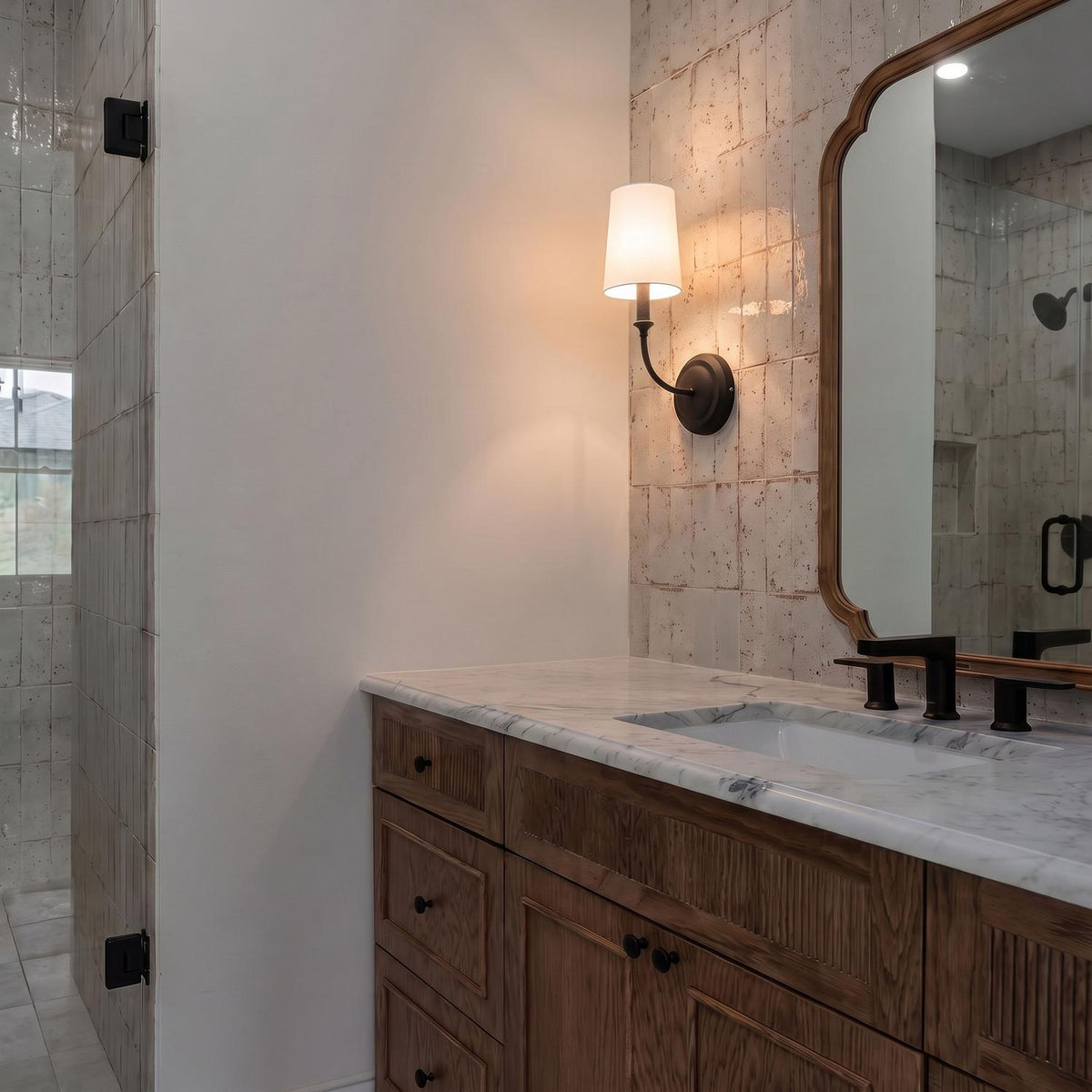 A transitional bathroom with the Estelle Wall Sconce 16", Forged Black and White Linen mounted on a beige tile wall beside a wood mirror and marble vanity.