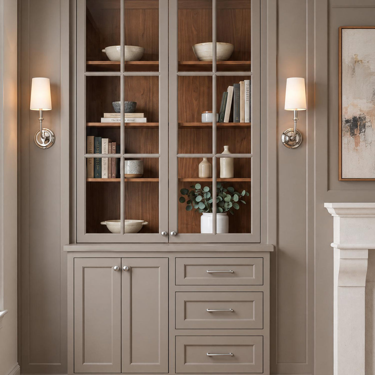 A transitional living room with Estelle Wall Sconce 16", Polished Nickel and White Linen fixtures flanking taupe glass-front cabinetry beside a carved stone fireplace.