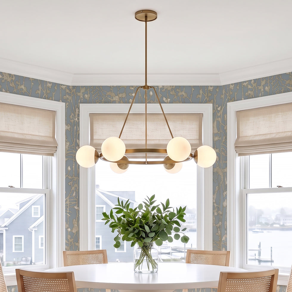 The Castell 6 Light Round Chandelier in Aged Brass with Opal Glass Globes is shown in a bright dining room with blue and tan wallapper and light oak wood chairs. Save 40% now during our Summer Sale.