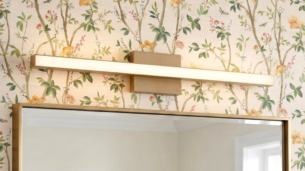 A Garrison LED Vanity Light 30", Satin Brass finish shown above a vanity mirror with colorful floral wallpaper.