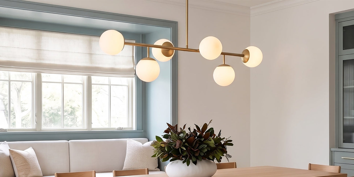 The Castell 6 Light Linear Chandelier in Aged Brass with Opal Glass Globes is shown in a bright dining room with blue accents and light oak wood. Save 40% now during our Summer Sale.