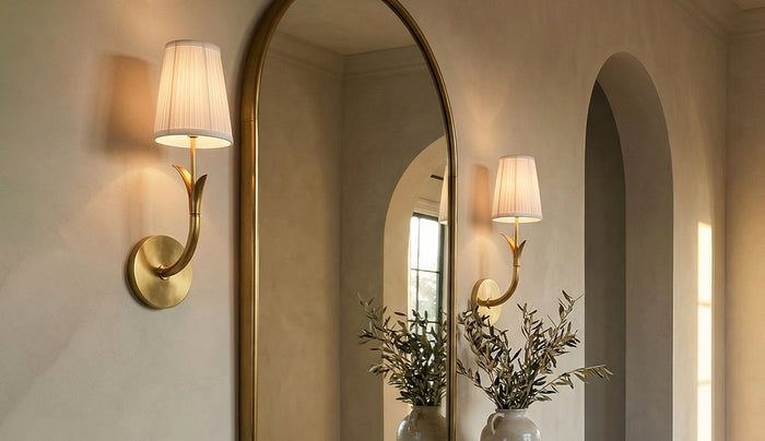 Two brass Magnolia wall sconces with a cream gathered-fabric drum shades, mounted on a textured beige plaster wall next to a large arched mirror. The mirror reflects the sconce and a vase of greenery, creating a symmetrical, elegant interior scene.