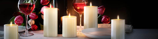 Group of ivory Infinity Wick flameless LED pillar candles, glowing Valentine's Day candles, on a romantic tabletop with pink roses and red wine.