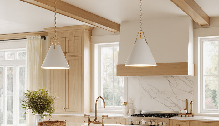 Two white metal Bowie 16" cone-shaped pendant lights with gold-toned hardware hang over a modern kitchen with light wood cabinets, a white marble backsplash, and a large white range hood.