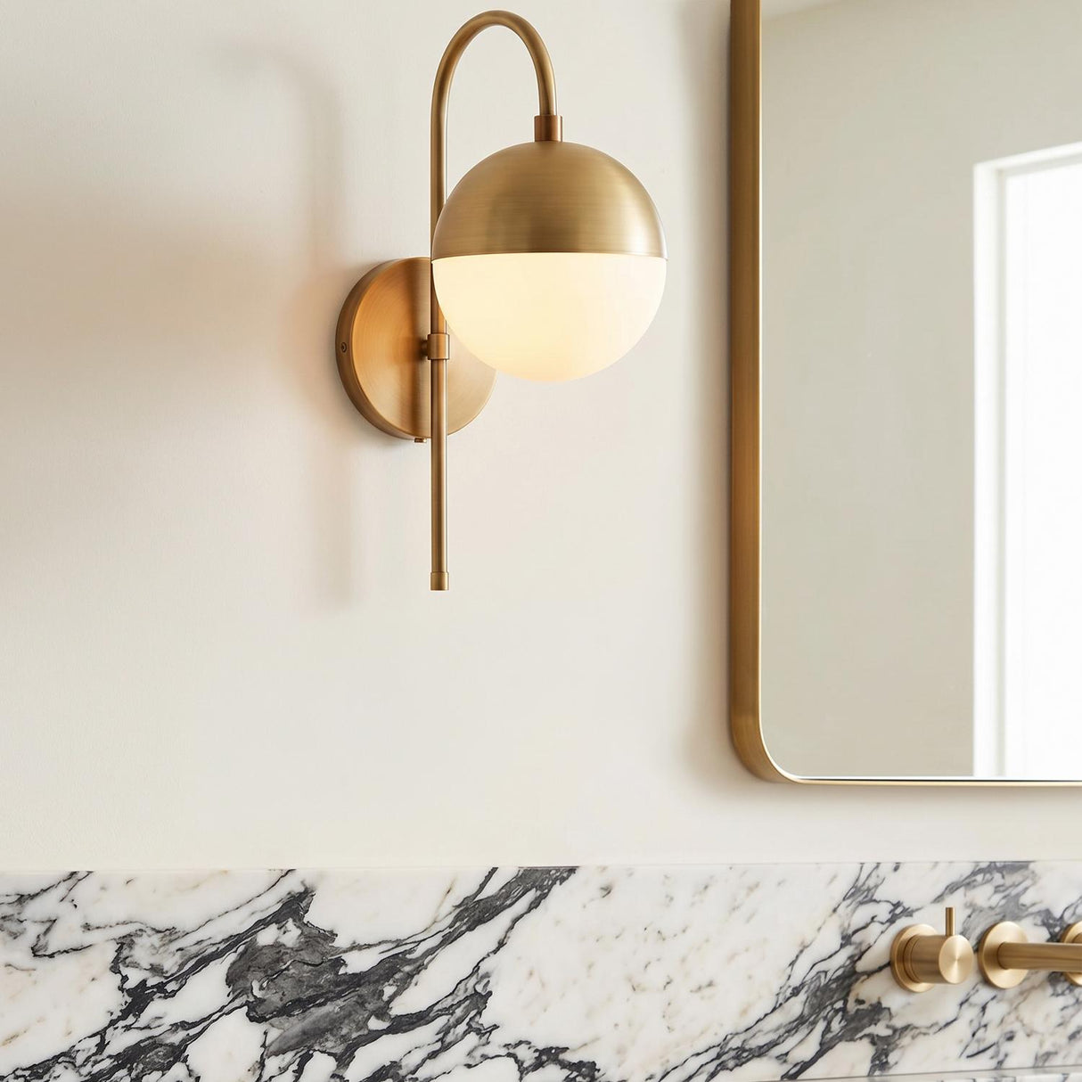 A contemporary bathroom featuring a Powell Wall Sconce with Hooded White Globe in aged brass mounted beside a mirror over a heavily veined marble backsplash.