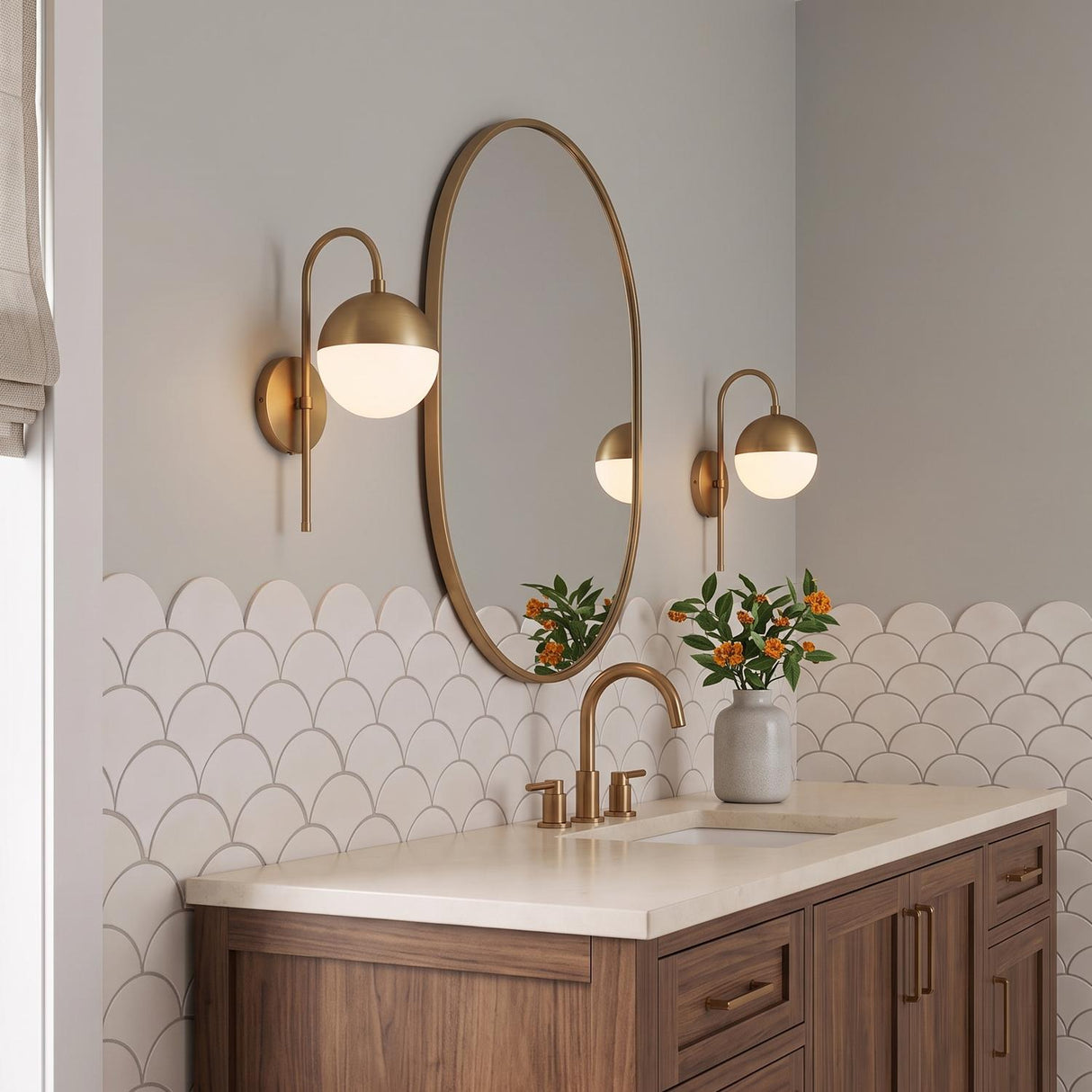 A contemporary bathroom with two Powell Wall Sconces with Hooded White Globes in aged brass flanking an oval mirror above a walnut vanity and scalloped backsplash.