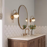 A contemporary bathroom with two Powell Wall Sconces with Hooded White Globes in aged brass flanking an oval mirror above a walnut vanity and scalloped backsplash.