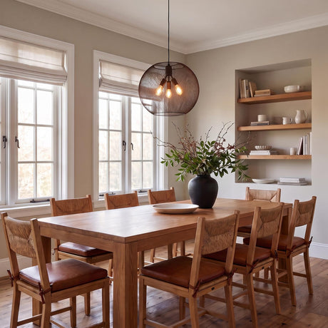 A transitional dining room with a bronze mesh globe pendant hanging over a natural wood table with woven-back chairs beside a built-in recessed bookshelf.