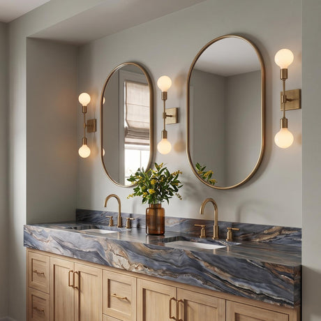 A modern bathroom with three Prospect 2 Light Wall Sconce, Aged Brass fixtures with frosted globe bulbs flanking two oval mirrors above a blue marble countertop.