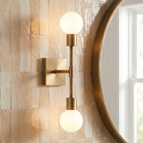 A contemporary bathroom with a Prospect 2 Light Wall Sconce, Aged Brass, featuring frosted glass globes mounted on peach-toned vertical tile beside a round mirror.