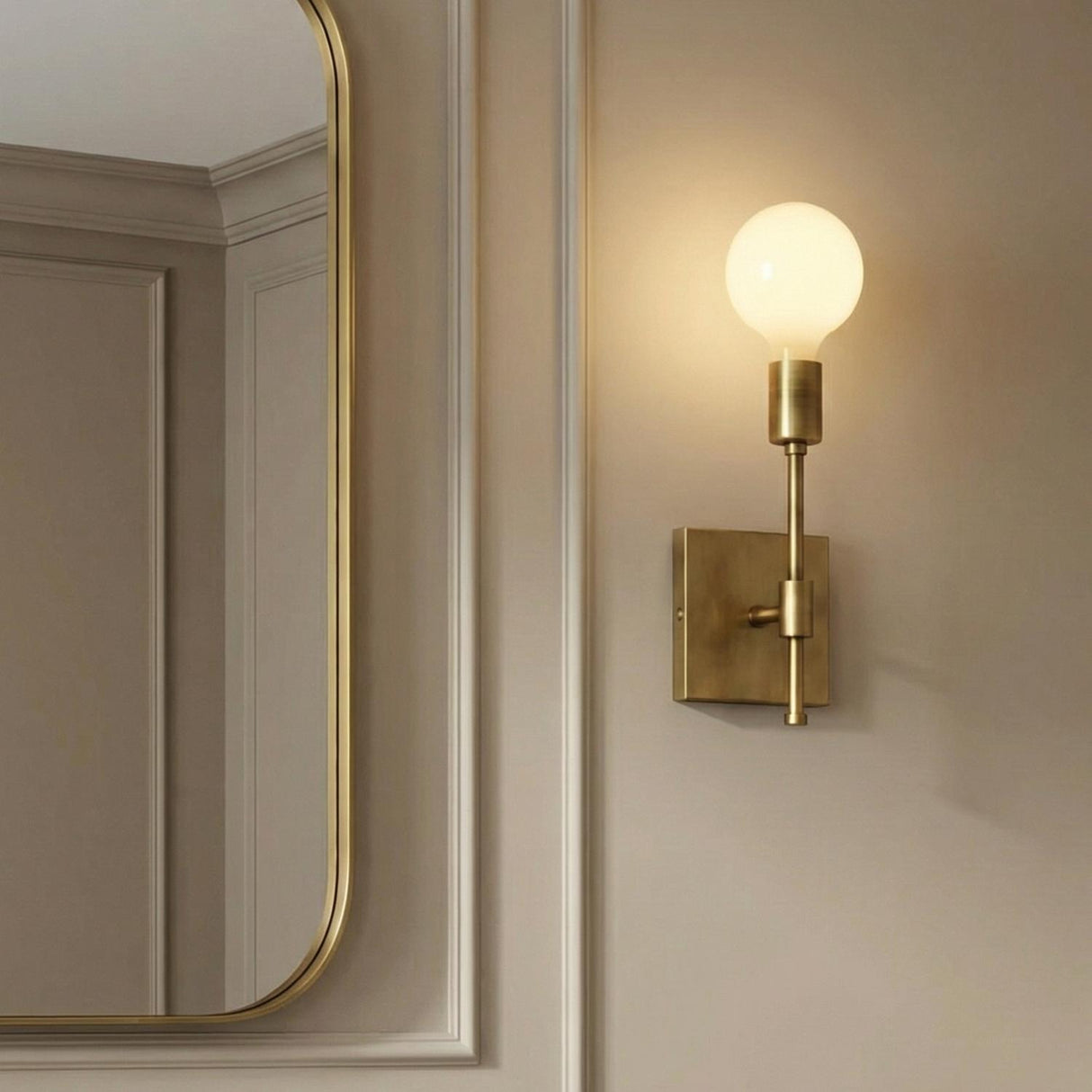 A transitional bathroom with the Prospect Wall Sconce, Aged Brass and an exposed globe bulb mounted next to a gold framed mirror on beige paneled walls.