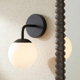 A modern bathroom with the Castell Single Globe Vanity Light, Matte Black featuring a frosted glass globe mounted on a beige textured wall beside a dark bobbin mirror.