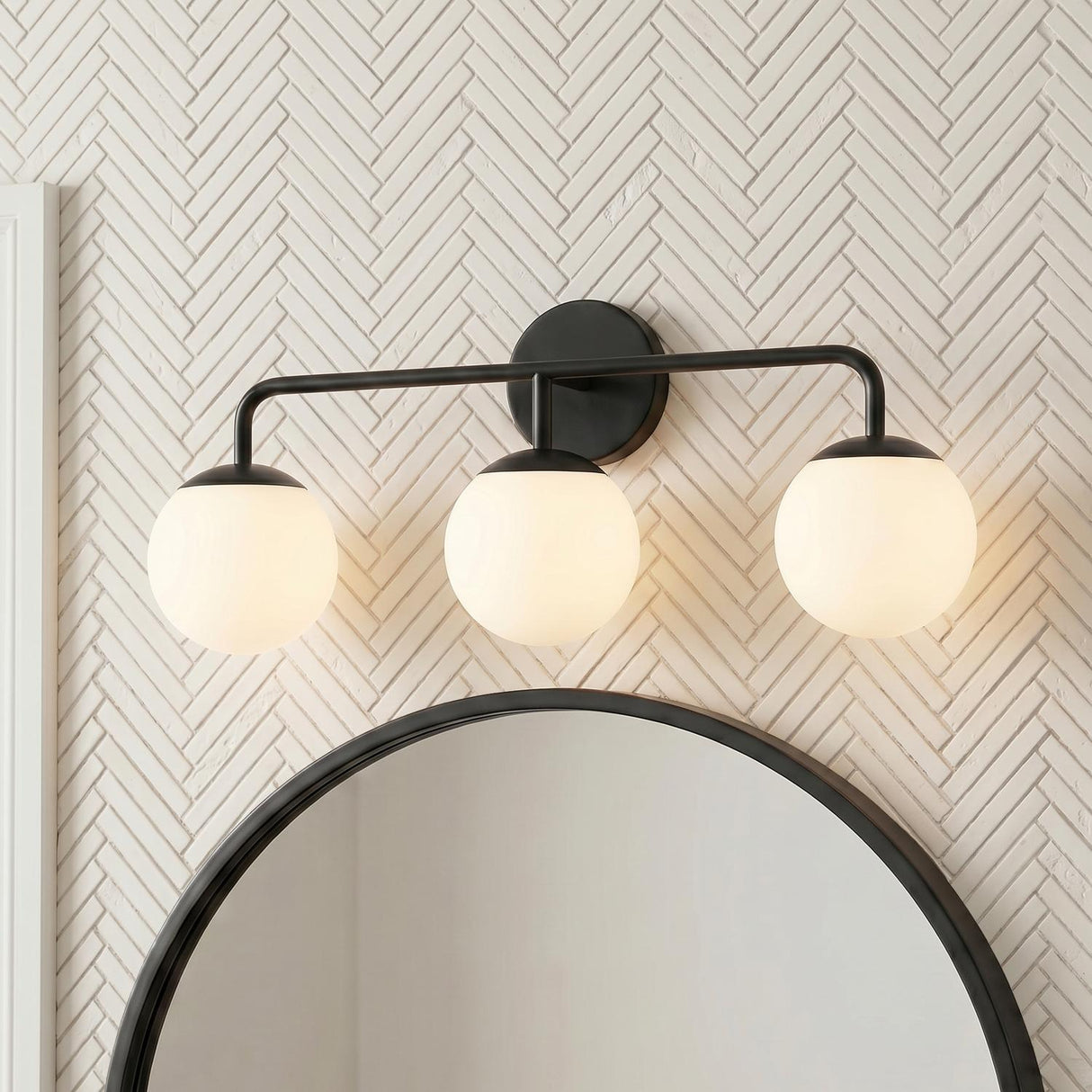 A modern bathroom features a Castell 3 Globe Vanity, Matte Black with frosted glass globes mounted above a round mirror on a white herringbone tile wall.