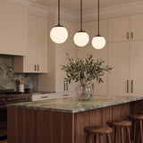A modern kitchen with three matte black globe pendants over a walnut island with a green marble countertop and matching backsplash with beige cabinets.