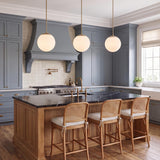 A transitional kitchen featuring three Castell 12" Globe Pendants in aged brass over a black marble island with blue cabinets and woven rattan stools.
