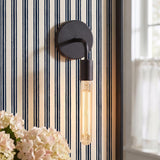 A coastal bedroom with a matte black Prospect Curved Wall Sconce with an exposed filament bulb mounted on blue striped wallpaper beside a black framed mirror.