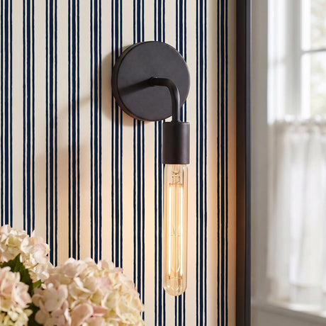 A coastal bedroom with a matte black Prospect Curved Wall Sconce with an exposed filament bulb mounted on blue striped wallpaper beside a black framed mirror.