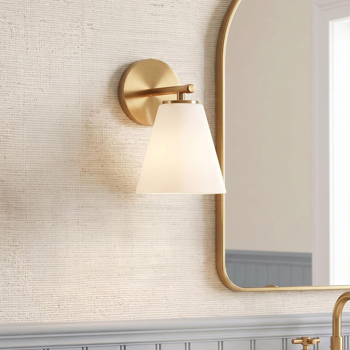 A transitional bathroom with a Carlisle Vanity Wall Sconce, Brushed Brass with Opal Glass, mounted on textured wallpaper beside a gold-framed arched mirror and gray wainscoting.