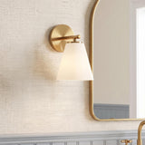A transitional bathroom with a Carlisle Vanity Wall Sconce, Brushed Brass with Opal Glass, mounted on textured wallpaper beside a gold-framed arched mirror and gray wainscoting.