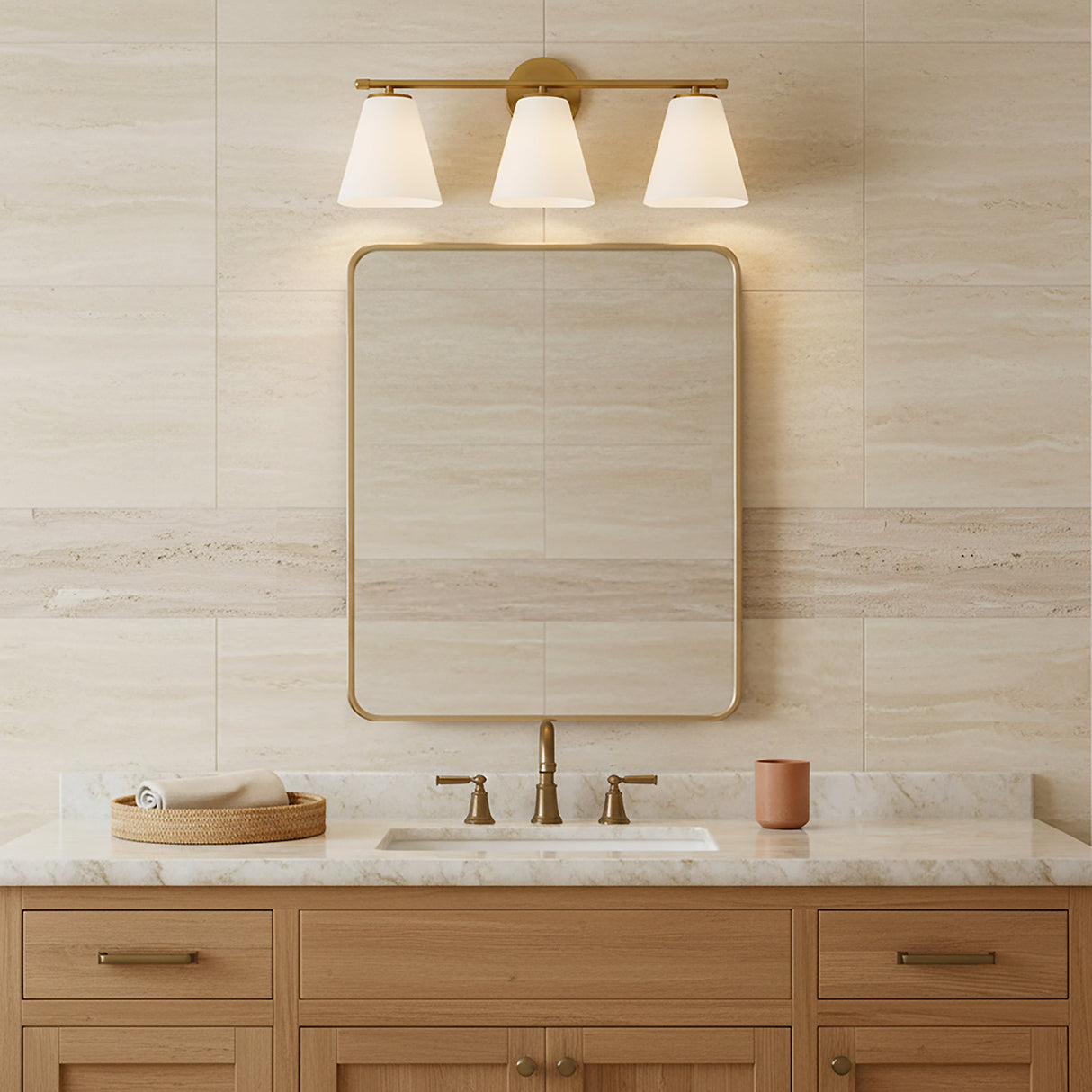 Carlisle 3 Light Vanity, Brushed Brass with Opal Glass - A vanity light fixture with three opal glass shades in a brushed brass finish is mounted above a framed mirror in a bathroom with travertine-style tile.