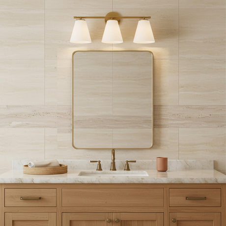 Carlisle 3 Light Vanity, Brushed Brass with Opal Glass - A vanity light fixture with three opal glass shades in a brushed brass finish is mounted above a framed mirror in a bathroom with travertine-style tile.