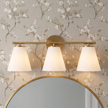 A transitional bathroom with the Carlisle 3 Light Vanity, Brushed Brass with Opal Glass, mounted above a round brass-framed mirror against floral patterned wallpaper.