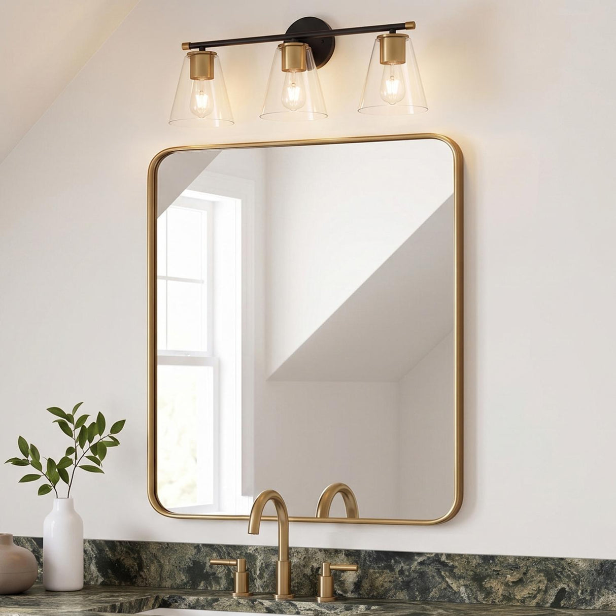 A transitional bathroom featuring the Carlisle 3 Light Vanity in matte black and brushed brass above a gold-framed mirror and green marble countertops under a sloped ceiling.
