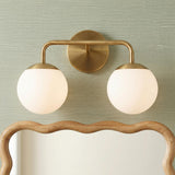 A modern bathroom with the Castell 2 Globe Vanity Light, Aged Brass featuring opal glass globes mounted above a wavy wood-framed mirror on sage green textured wallpaper.