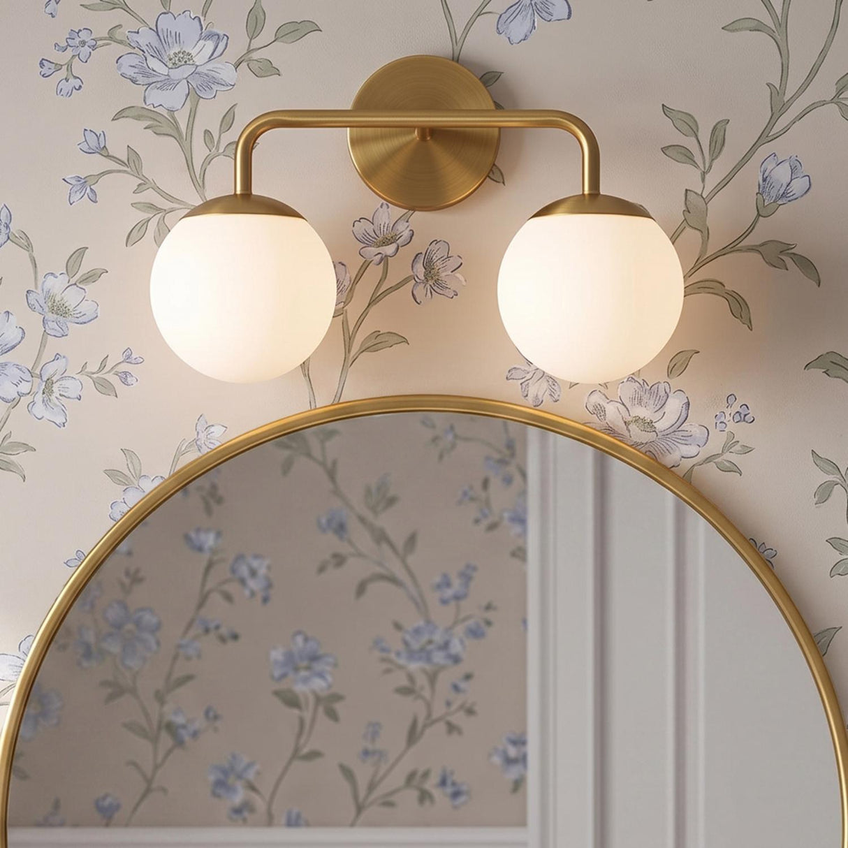 A transitional bathroom with the Castell 2 Globe Vanity Light, Aged Brass mounted above a round gold-rimmed mirror on floral wallpaper with white wall paneling.