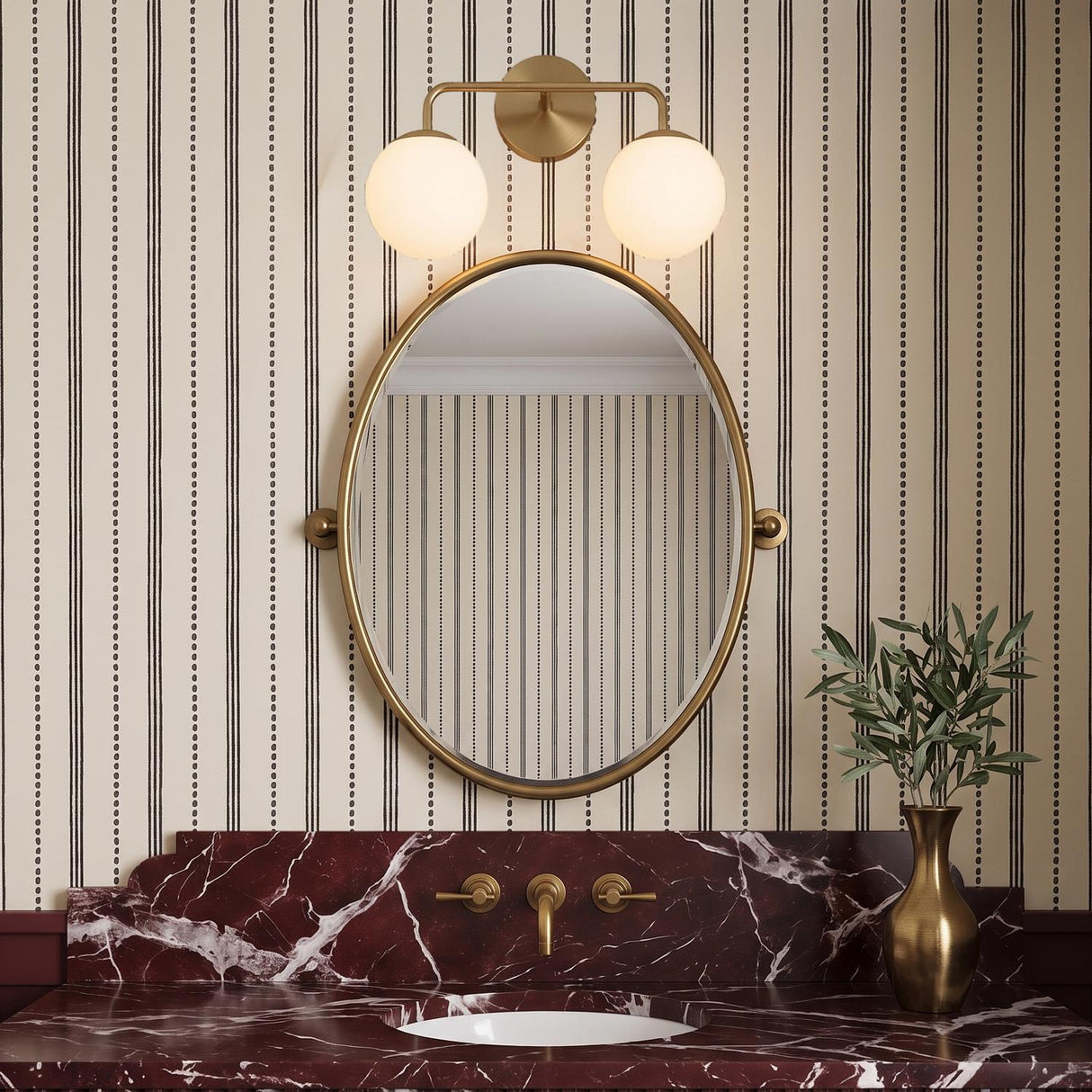 A transitional bathroom featuring the Castell 2 Globe Vanity Light, Aged Brass, mounted above an oval mirror and a deep red veined marble vanity with striped wallpaper.