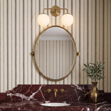 A transitional bathroom featuring the Castell 2 Globe Vanity Light, Aged Brass, mounted above an oval mirror and a deep red veined marble vanity with striped wallpaper.