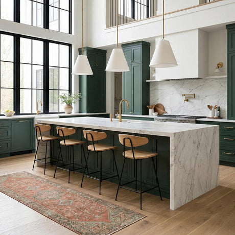 A double-height kitchen features three Luca Small 13" Conical Pendant, Satin Brass fixtures with white linen shades hanging over a marble waterfall island and green cabinets.